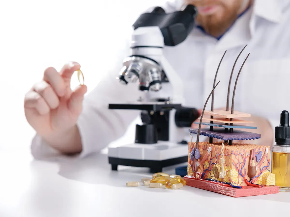 Stock photograph of a model: a researcher at a microscope holding a capsule, beside a model of the layers of the skin