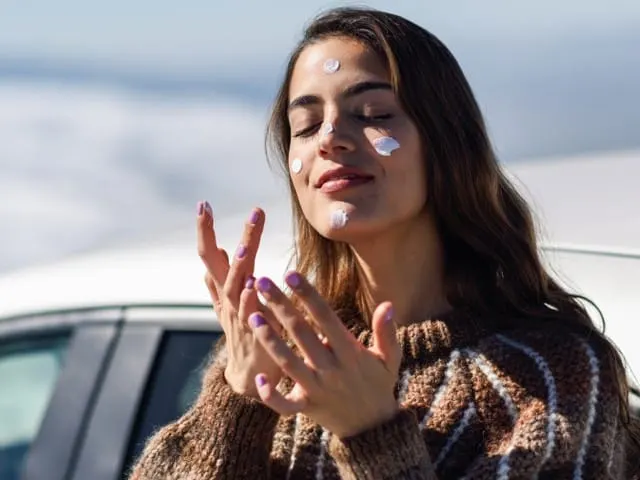 Stock photograph of a model: a woman in a brown sweater with dabs of sunscreen on her face, eyes closed, in front of a car on a sunny day