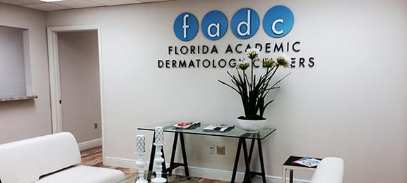 The Florida Academic Dermatology Center reception in Coral Gables: the fadc sign on a pale wall above a glass desk with white flowers and white armchairs