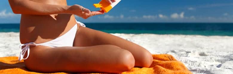 Stock photograph of a model: a woman in a white bikini sitting on a beach towel, holding a bottle of sunscreen