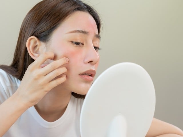 Stock photograph of a model: a young woman with reddened cheeks touching her face while looking into a hand mirror