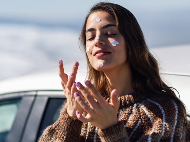 Stock photograph of a model: a woman in a brown sweater with dabs of sunscreen on her face, eyes closed, in front of a car on a sunny day