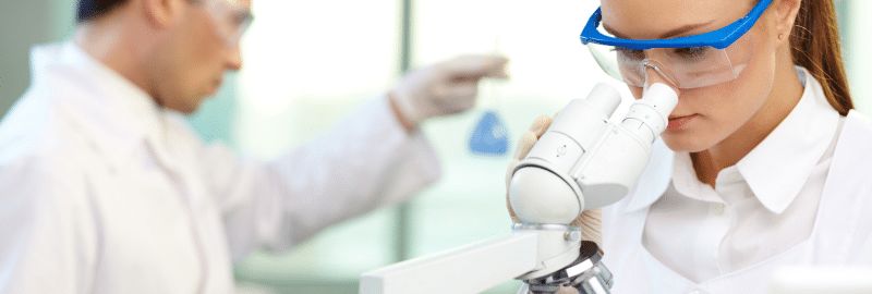 Stock photograph of models: a woman looking through a microscope while a colleague holds a test tube in a lab