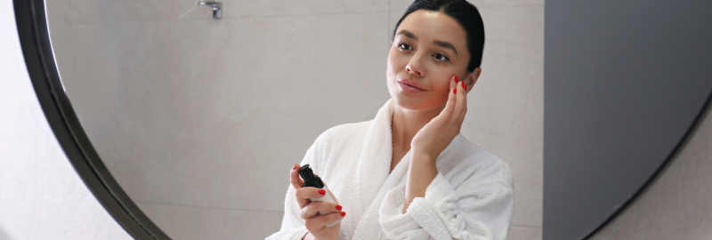Stock photograph of a model: a woman in a white robe applying skincare product to her face in a bathroom mirror