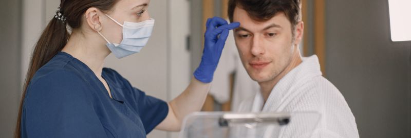 Stock photograph of models: a masked clinician in blue scrubs and gloves examining a man's forehead